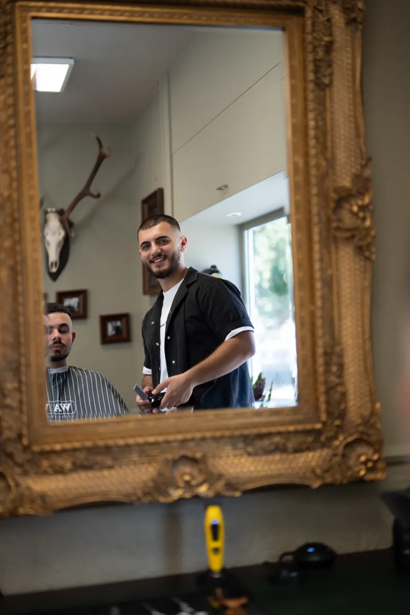 Lorik, Barbier, lächelt hinter seinem Kunden im Goldspiegel